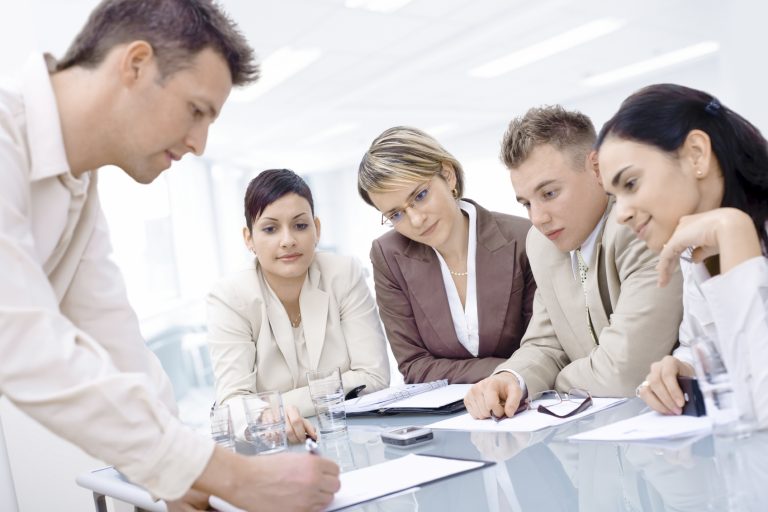 4 people sitting at a desk looking at a fifth person who is writting on a piece of paper.