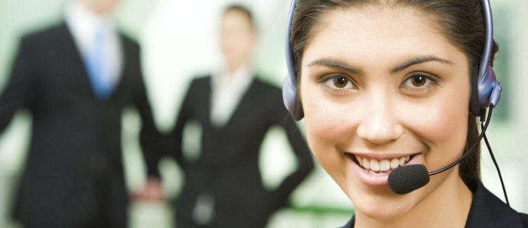 A person with a microphone headset on, with two people in the background in suits blurred out.